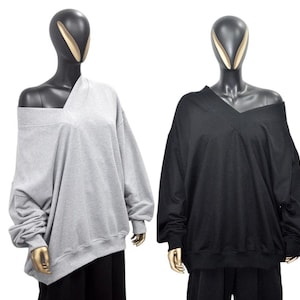 Könnte beinhalten: Zwei Off-Shoulder-Sweatshirts auf Schaufensterpuppen. Eines ist hellgrau, das andere ist schwarz. Beide haben einen weiten V-Ausschnitt und lange Ärmel. Die Sweatshirts werden auf schwarzen Schaufensterpuppen mit goldenen Akzenten präsentiert.