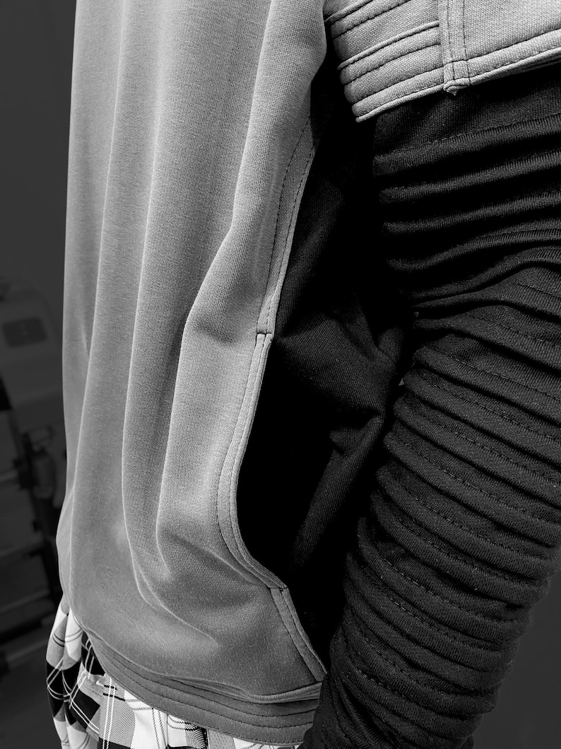 K&ouml;nnte beinhalten: Ein grauer Kapuzenpullover mit einer K&auml;ngurutasche und einem schwarzen gerippten &Auml;rmel. Der Kapuzenpullover hat eine Kapuze mit Kordelzug und einen kontrastierenden schwarzen Einsatz. Der untere Teil des Kapuzenpullovers ist sichtbar, mit einem Karomuster.