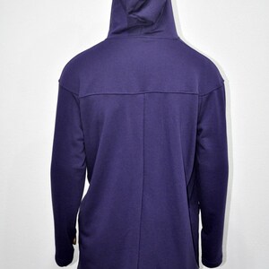 Puede incluir: Una sudadera con capucha de color p&uacute;rpura oscuro con capucha y mangas largas. La parte trasera de la sudadera con capucha es visible, mostrando un detalle de costura en la parte superior de la espalda. La parte inferior de la sudadera con capucha es visible, con pantalones de ch&aacute;ndal grises.