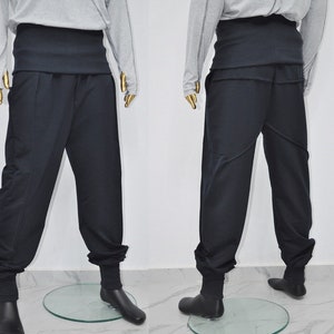 May include: Two mannequins display black pants with a high, wide waistband and fitted ankles. The pants have a relaxed fit through the legs. The mannequins are wearing gray long-sleeved shirts. The pants are displayed on clear, circular platforms.