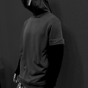 K&ouml;nnte beinhalten: Schwarz-Wei&szlig;-Bild einer Person in einem schwarzen Kapuzenpullover mit einem einzigartigen, geschichteten Kapuzendesign. Die &Auml;rmel sind lang und strukturiert. Die Person tr&auml;gt auch eine schwarz-wei&szlig; karierte Hose. Der Hintergrund ist ein dunkles Grau.