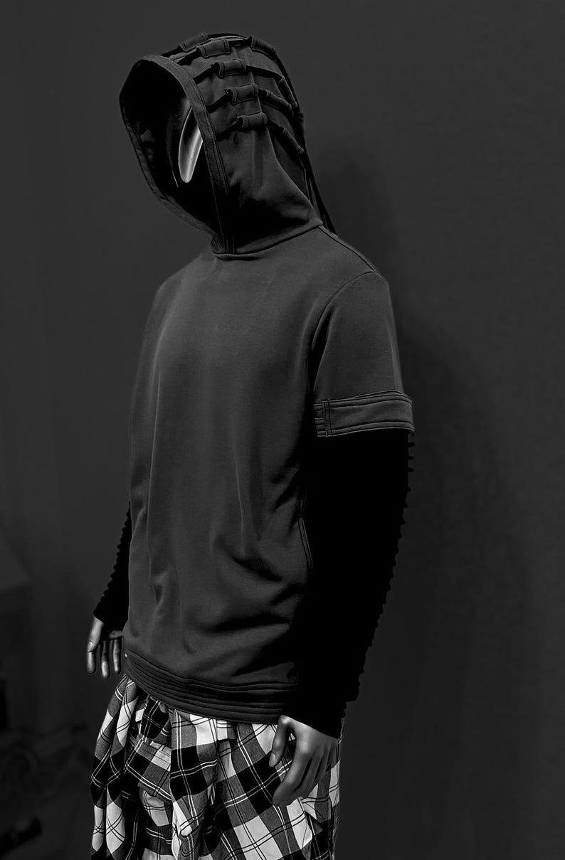 K&ouml;nnte beinhalten: Schwarz-Wei&szlig;-Bild einer Person in einem schwarzen Kapuzenpullover mit einem einzigartigen, geschichteten Kapuzendesign. Die &Auml;rmel sind lang und strukturiert. Die Person tr&auml;gt auch eine schwarz-wei&szlig; karierte Hose. Der Hintergrund ist ein dunkles Grau.
