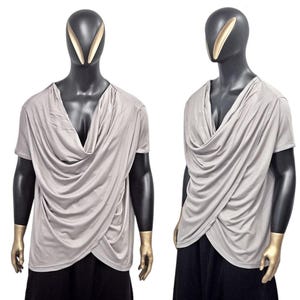 May include: Two mannequins display a light gray draped-front top with short sleeves. The fabric wraps across the front, creating a layered effect. The mannequins have black bodies and gold accents on their faces and hands.