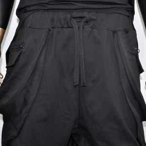 以下が含まれることがあります： 黒のドロップクロッチパンツ。ウエストはドローストリング、大きなドレープポケット付き。リラックスフィットで、両側にジッパーポケットが付いています。ゴールドカラーの手のマネキンに展示されています。