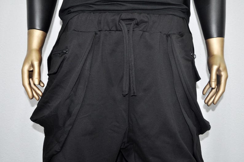 以下が含まれることがあります： 黒のドロップクロッチパンツ。ウエストはドローストリング、大きなドレープポケット付き。リラックスフィットで、両側にジッパーポケットが付いています。ゴールドカラーの手のマネキンに展示されています。