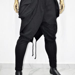 以下が含まれることがあります： ユニークなドレープデザインの黒いアバンギャルドパンツ。ハイウエスト、ジッパーポケット、ドローストリングが特徴です。下腿はフィットし、独特のシルエットを作り出しています。マネキンに展示されています。