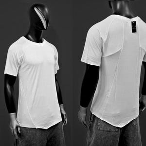 Puede incluir: Una camiseta blanca de manga corta con un dobladillo asimétrico único se muestra en un maniquí. La camiseta tiene cuello redondo y un diseño minimalista. El maniquí es negro y el fondo es gris oscuro.