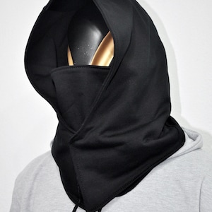 May include: Black balaclava hood with an integrated face covering. The hood is made of a soft, dark fabric and has a drawstring closure. The balaclava is designed to cover the head, neck, and face.