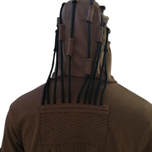 K&ouml;nnte beinhalten: Ein brauner Kapuzenpullover mit schwarzen Kordeln und Akzenten. Die Kapuze hat mehrere schwarze Kordeln mit braunen zylindrischen Akzenten. Auf der R&uuml;ckseite des Sweatshirts befindet sich ein gesticktes geometrisches Design und vertikale N&auml;hte.