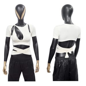 Puede incluir: Un top blanco corto con un diseño cruzado en la parte delantera y trasera. El top tiene mangas cortas y un escote redondo.