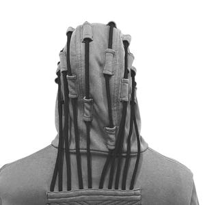K&ouml;nnte beinhalten: Grauer Kapuzenpullover mit einem einzigartigen Design mit schwarzen Kordeln, die sich von der Kapuze erstrecken. Die R&uuml;ckseite des Kapuzenpullovers hat eine Tasche mit einem geometrischen Muster. Das Kleidungsst&uuml;ck ist aus weichem Material.