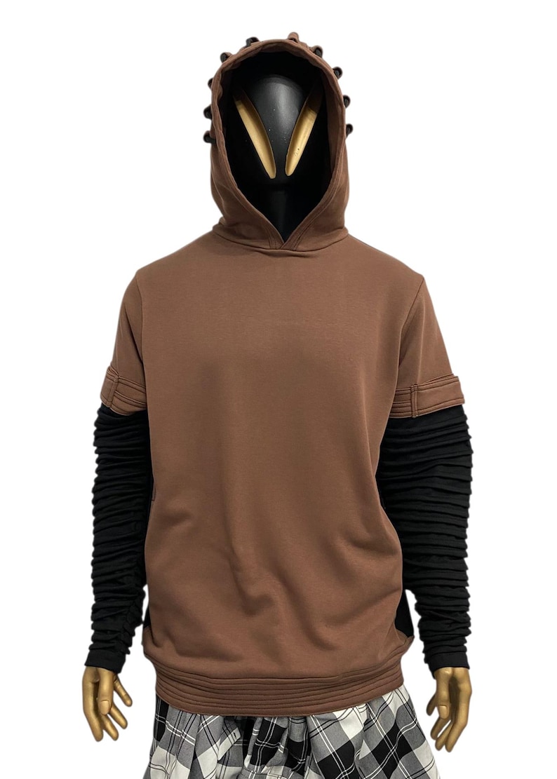 K&ouml;nnte beinhalten: Brauner Kapuzenpullover mit schwarzer Kapuze und &Auml;rmeln. Der Kapuzenpullover hat ein einzigartiges Design mit strukturierter Kapuze und &Auml;rmeln. Die &Auml;rmel sind lang und eng anliegend, und der Kapuzenpullover ist aus einem weichen Material gefertigt. Die Schaufensterpuppe tr&auml;gt einen karierten Rock.