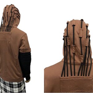 K&ouml;nnte beinhalten: Brauner Kapuzenpullover mit schwarzen Akzenten. Die Kapuze hat mehrere schwarze Kordeln, und die &Auml;rmel sind schwarz mit gerippten Details. Die R&uuml;ckseite des Sweatshirts hat ein strukturiertes Design. Die Person tr&auml;gt ein schwarz-wei&szlig;es Karomuster.