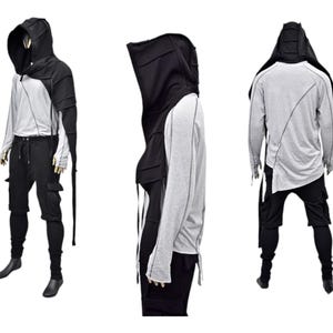 Könnte beinhalten: Ein schwarzes Kapuzenkleidungsstück mit einem langen, fließenden Umhang. Das Kleidungsstück hat ein einzigartiges Design mit mehreren Schichten und Riemen. Das Model trägt das Kleidungsstück über einem grauen Langarmshirt und einer schwarzen Hose.