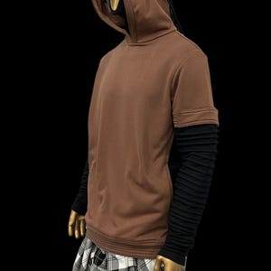 K&ouml;nnte beinhalten: Brauner Kapuzenpullover mit schwarzen Akzenten und langen schwarzen &Auml;rmeln. Die Kapuze hat ein einzigartiges Design mit schwarzen Kordeln. Die Schaufensterpuppe tr&auml;gt einen schwarz-wei&szlig; karierten Rock. Das Outfit wird vor schwarzem Hintergrund pr&auml;sentiert.