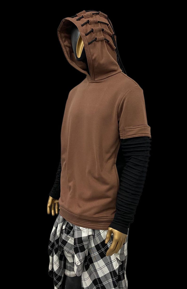 K&ouml;nnte beinhalten: Brauner Kapuzenpullover mit schwarzen Akzenten und langen schwarzen &Auml;rmeln. Die Kapuze hat ein einzigartiges Design mit schwarzen Kordeln. Die Schaufensterpuppe tr&auml;gt einen schwarz-wei&szlig; karierten Rock. Das Outfit wird vor schwarzem Hintergrund pr&auml;sentiert.