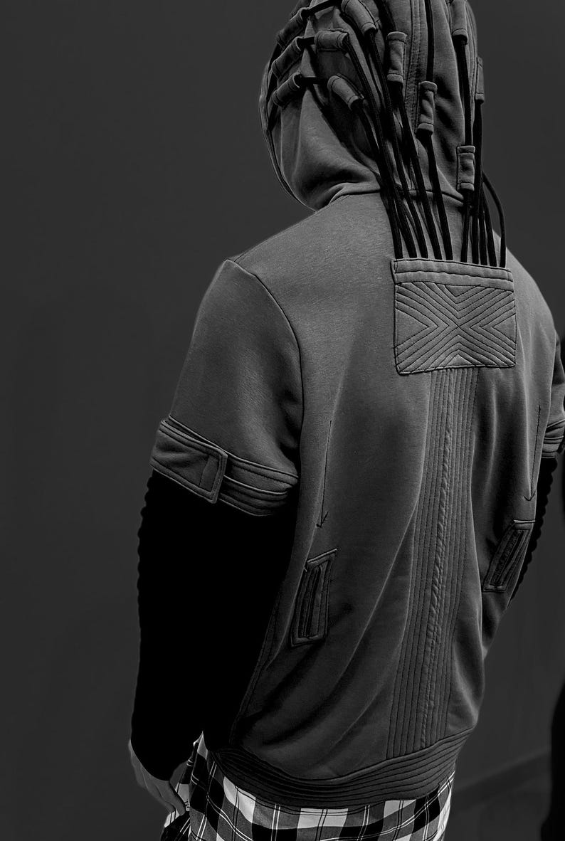 K&ouml;nnte beinhalten: Ein grauer Kapuzenpullover mit einem einzigartigen Design. Die R&uuml;ckseite hat ein strukturiertes Panel mit einem X-f&ouml;rmigen Muster und vertikalen N&auml;hten. Die Kapuze hat mehrere schwarze Kordeln. Die &Auml;rmel haben eine schwarze Rippstruktur. Der untere Teil ist ein Karomuster.