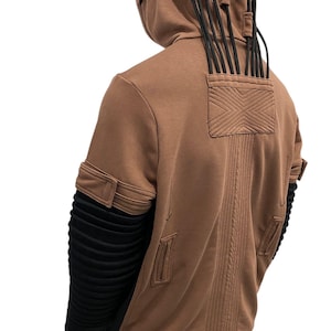 K&ouml;nnte beinhalten: Brauner Kapuzenpullover mit schwarzen Akzenten. Er hat eine strukturierte R&uuml;ckseite, eine Tasche mit X-Design und schwarze Kordeln, die von der Kapuze ausgehen. Die &Auml;rmel haben schwarzen, gerippten Stoff und braune Manschetten.