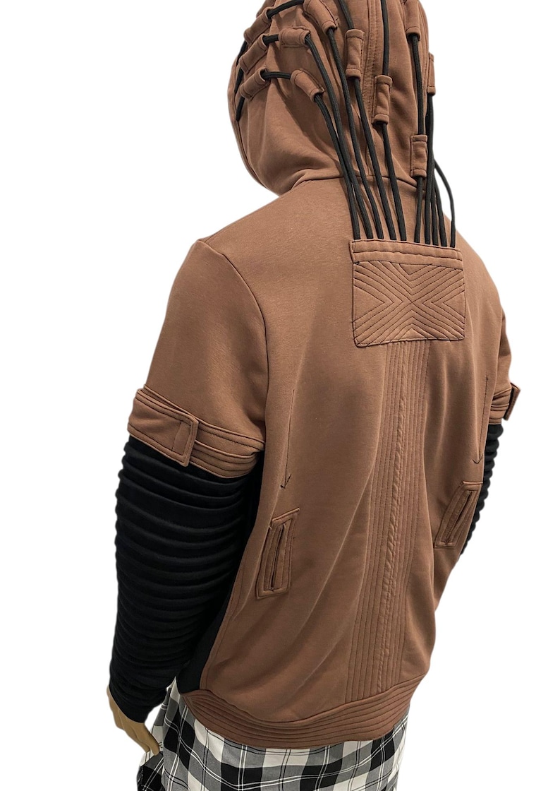 K&ouml;nnte beinhalten: Brauner Kapuzenpullover mit schwarzen Akzenten. Er hat eine strukturierte R&uuml;ckseite, eine Tasche mit X-Design und schwarze Kordeln, die von der Kapuze ausgehen. Die &Auml;rmel haben schwarzen, gerippten Stoff und braune Manschetten.