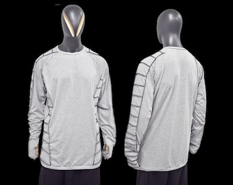 Puede incluir: Una camiseta de manga larga gris claro con detalles de costuras en gris oscuro. La camiseta se muestra en un maniquí, mostrando las vistas delantera y trasera. Las costuras crean un patrón geométrico en las mangas y los laterales.