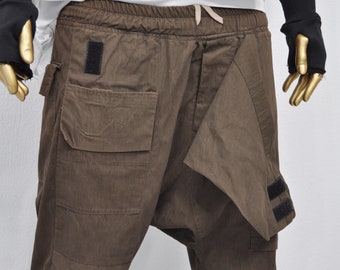 May include: Brown cargo pants with a unique design featuring a flap that wraps around the front. The pants have a drawstring waistband and multiple pockets.
