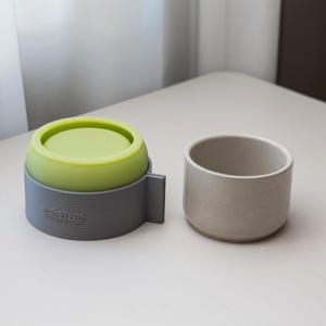 May include: A grey mould with a lime green insert next to a small, light grey ceramic cup. The mould has the word "archetype" printed on the side. The cup is cylindrical with a smooth, matte finish.
