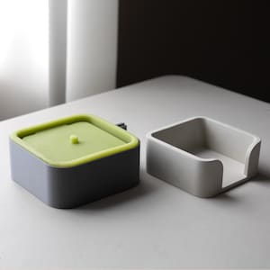 May include: Two contemporary, square containers. One is grey with a lime green lid and a small knob. The other is a simple, open, white container. Both are on a white surface.