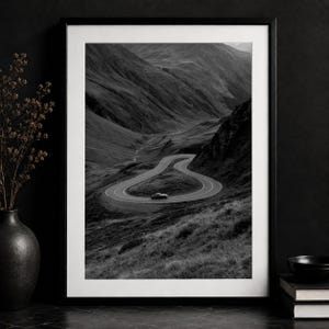 Puede incluir: Una impresión de arte mural enmarcada en blanco y negro que muestra una carretera de montaña sinuosa con un solo vehículo tomando una curva cerrada. El paisaje presenta laderas escarpadas y texturizadas en tonos monocromáticos de alto contraste.
