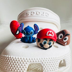 May include: White Crocs shoe with four cartoon character charms: a red Spider-Man, a blue Stitch, a red-hatted Mario, and a brown Goomba. The charms are attached to the top of the shoe.