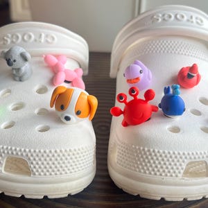 May include: White Crocs shoes decorated with colorful charms. One shoe features a koala, a pink balloon animal, a dog, and a green frog. The other shoe has a purple dolphin, a red crab, a blue whale, a red bird, a yellow duck, and a shark.