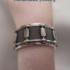 Puede incluir: Un anillo plateado con forma de tambor, con una superficie texturizada y detalles rectangulares. El anillo es artesanal y presenta el texto "SUNFASHIONHUB Handmade Jewelry".