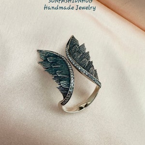 Puede incluir: Un anillo de plata con dos alas a cada lado. Las alas est&aacute;n detalladas con acentos negros y blancos. El anillo est&aacute; sobre un fondo rosa.