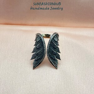 Puede incluir: Un anillo de plata con dos alas negras, cada ala est&aacute; detallada con un dise&ntilde;o de plumas y adornada con peque&ntilde;as piedras negras.