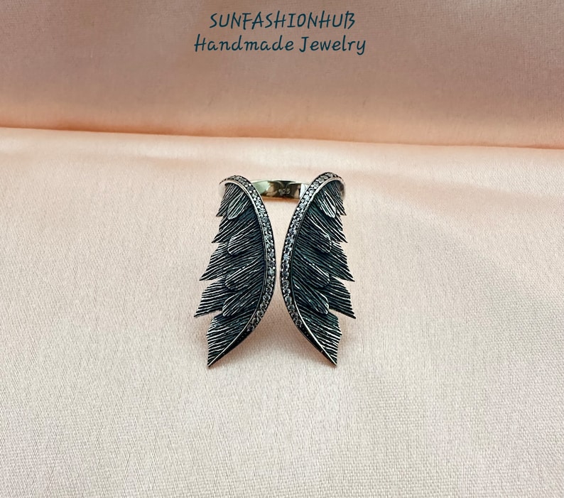 Puede incluir: Un anillo de plata con dos alas negras, cada ala est&aacute; detallada con un dise&ntilde;o de plumas y adornada con peque&ntilde;as piedras negras.