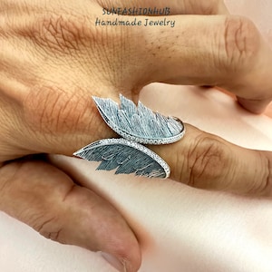 Puede incluir: Un anillo de plata con dos alas, cada ala est&aacute; detallada con un dise&ntilde;o de plumas texturizado. El anillo est&aacute; adornado con peque&ntilde;as piedras transparentes.