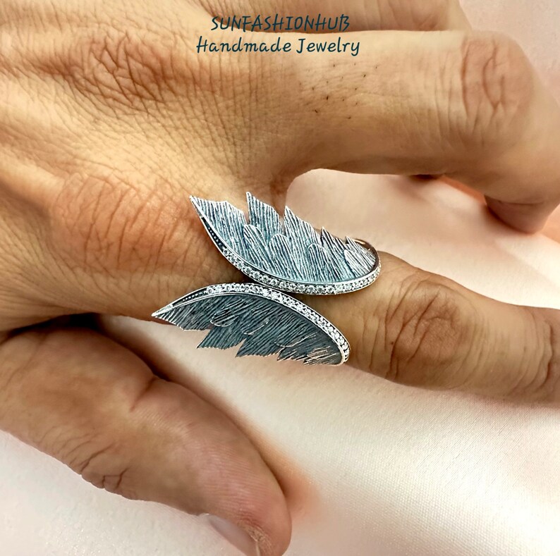 Puede incluir: Un anillo de plata con dos alas, cada ala est&aacute; detallada con un dise&ntilde;o de plumas texturizado. El anillo est&aacute; adornado con peque&ntilde;as piedras transparentes.