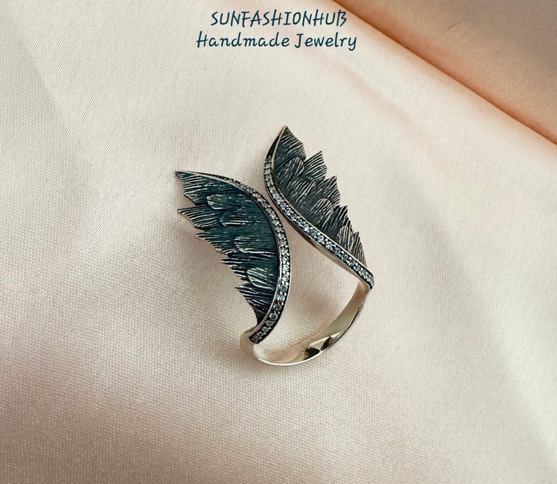 Puede incluir: Un anillo de plata con dos alas a cada lado. Las alas est&aacute;n detalladas con acentos negros y blancos. El anillo est&aacute; sobre un fondo rosa.