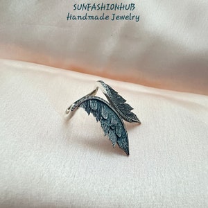 Puede incluir: Un anillo de plata con dos alas de plumas negras. El anillo est&aacute; sobre un fondo de tela rosa. SUNFASHIONHUB Joyer&iacute;a hecha a mano.