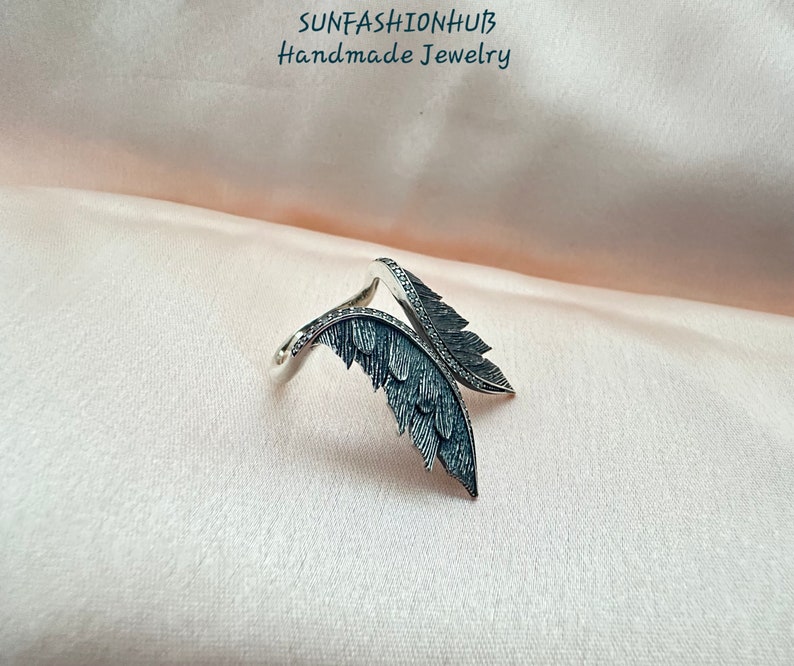 Puede incluir: Un anillo de plata con dos alas de plumas negras. El anillo est&aacute; sobre un fondo de tela rosa. SUNFASHIONHUB Joyer&iacute;a hecha a mano.