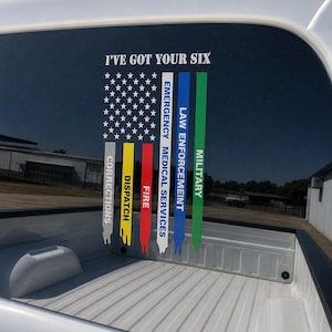 May include: A vinyl decal on a truck window featuring a stylized American flag with white stars on a black field. Below, vertical stripes in grey, yellow, red, white, blue, and green are labeled CORRECTIONS, DISPATCH, FIRE, EMERGENCY MEDICAL SERVICES, LAW ENFORCEMENT, and MILITARY. The text I'VE GOT YOUR SIX is printed above.