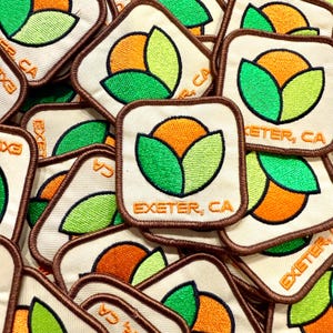 May include: A collection of square embroidered patches. Each patch features a stylized orange and green design with the text "EXETER, CA" in orange. The patches have a cream-colored background and a brown border. The design appears to be a stylized fruit or flower.