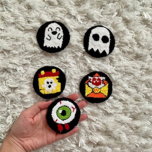 May include: Five Halloween-themed felt ornaments. The ornaments are round and feature a ghost, an eye, a ghost in a calendar, a ghost in an envelope, and a ghost with a smile. The ornaments are black with white and coloured details.