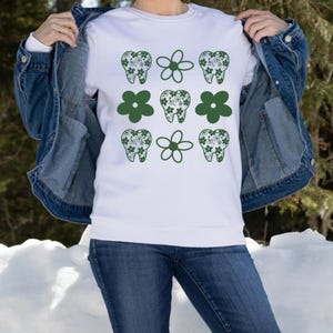 Pode incluir: Camisola branca com um padrão de dentes e flores verdes. Os dentes são preenchidos com um design floral. A camisola é usada com uma jaqueta jeans e jeans azuis. O fundo é uma cena externa com neve.