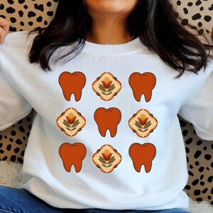 May include: White sweatshirt with a pattern of orange teeth and floral designs. The teeth are arranged in a grid pattern with a floral design in between each tooth.