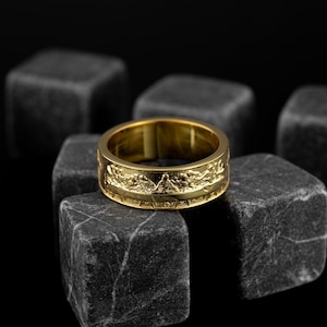 Pode incluir: Um anel de banda de ouro com um design texturizado. O anel está sobre uma superfície preta.