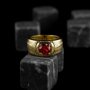 May include: A gold ring with a textured band and a red gemstone set in a square bezel.