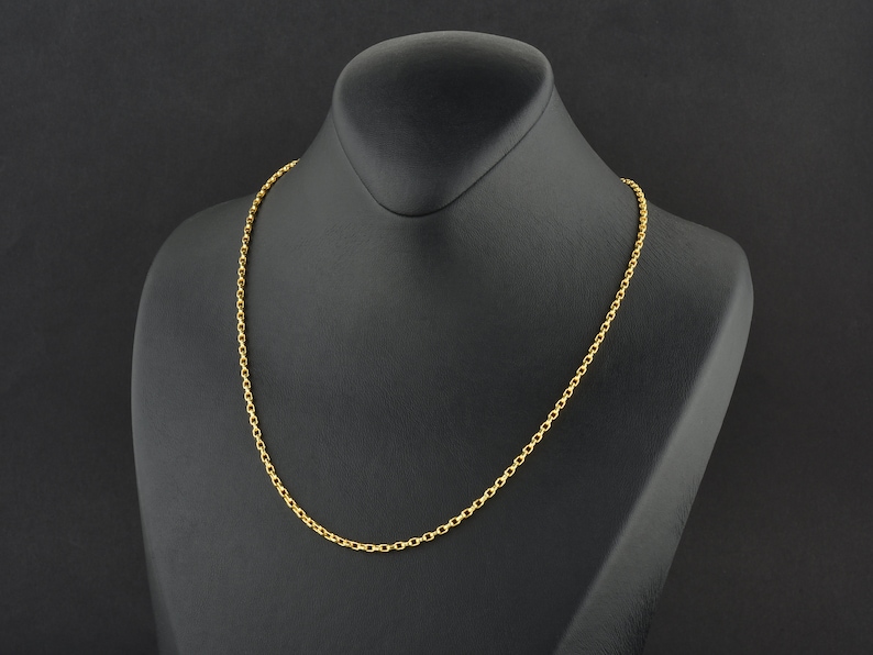 May include: A gold-colored chain necklace displayed on a black mannequin. The necklace is a simple, delicate design with small, evenly spaced links. The background is a solid black, highlighting the jewelry.