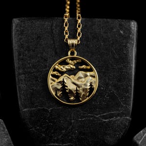 Könnte beinhalten: Eine goldene Halskette mit einem runden Anhänger, der eine detaillierte Silhouette von Bergen und Bäumen zeigt. Der Anhänger befindet sich auf einem schwarzen Hintergrund.