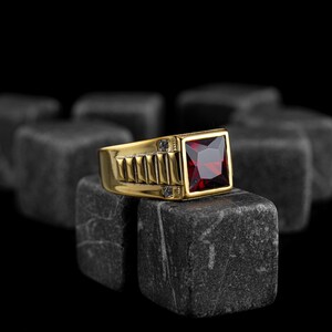 Könnte beinhalten: Ein goldener Ring mit einem quadratischen roten Edelstein und kleinen klaren Edelsteinen auf der Ringschiene. Der Ring liegt auf einem Stapel grauer Steine.