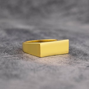 May include: A gold-coloured ring with a rectangular design. The ring has a flat top and a smooth, polished surface. The ring is set against a grey background.